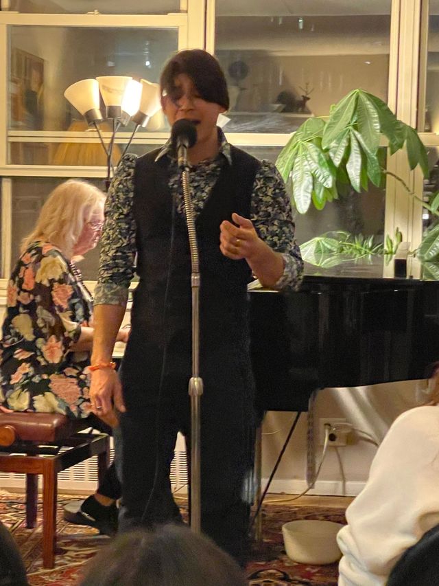Singer at a microphone in a room with a grand piano and seated accompanist.