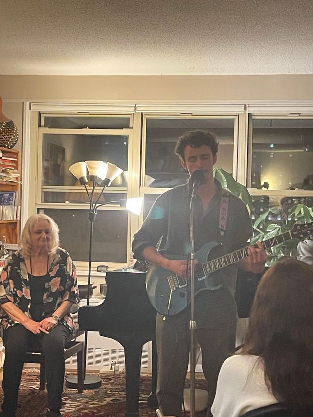 Musician singing and playing guitar at a small indoor gathering, with a seated audience nearby