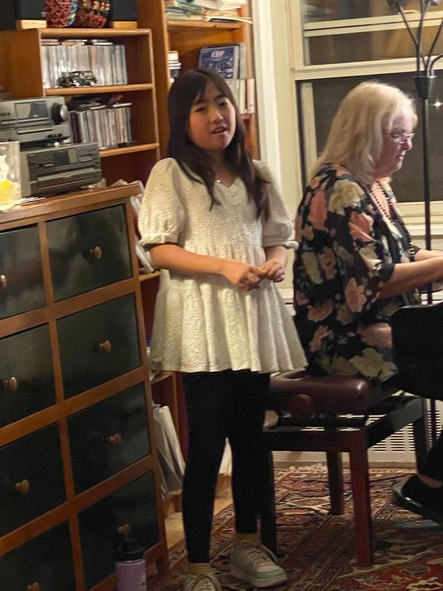 Girl in a white dress stands in a cozy room beside a seated woman near shelves and a chair.