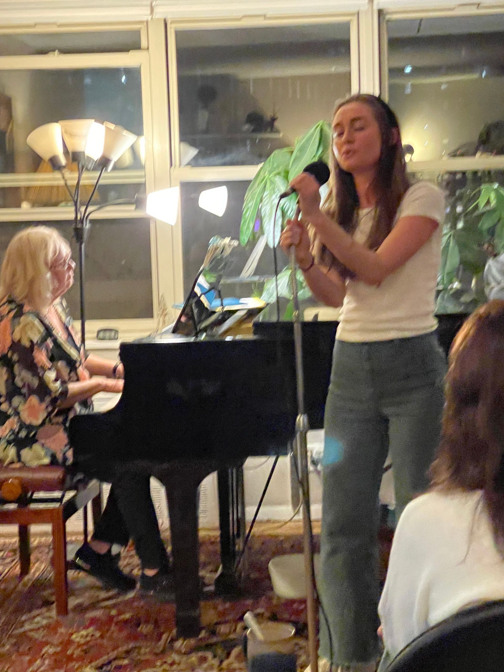 Singer performing beside a piano at a cozy indoor concert with audience nearby