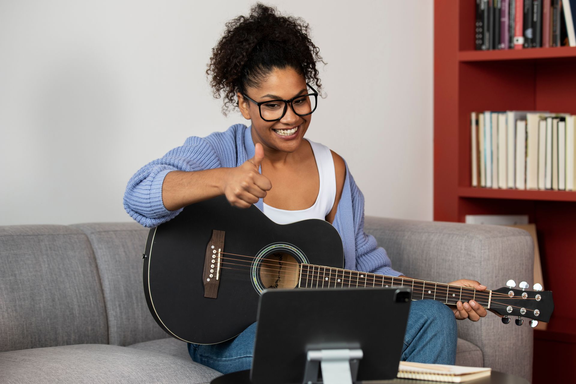 A woman is sitting on a couch playing an acoustic guitar and giving a thumbs up.