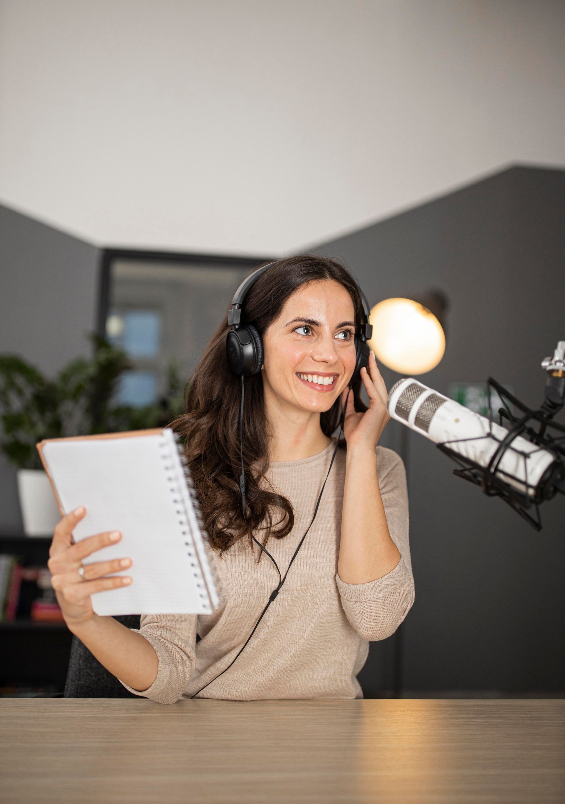 A woman wearing headphones is holding a notebook and talking into a microphone.