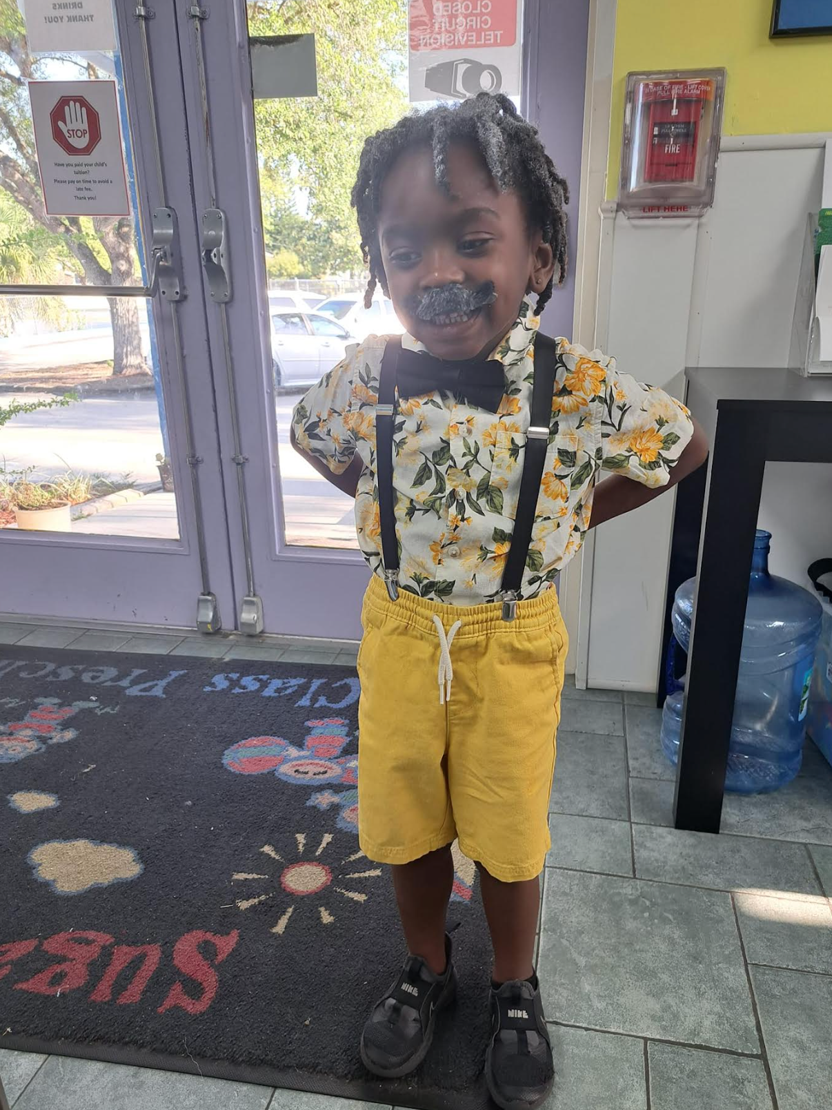 A young boy wearing a bow tie and suspenders is standing in front of a door.