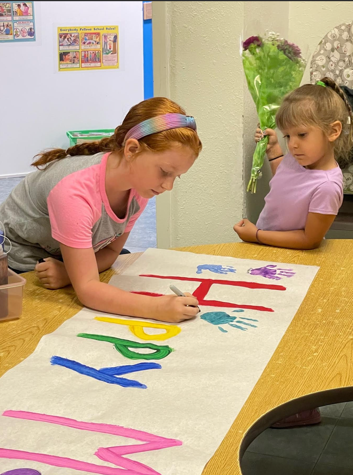Two young girls are sitting at a table painting a banner that says happy birthday