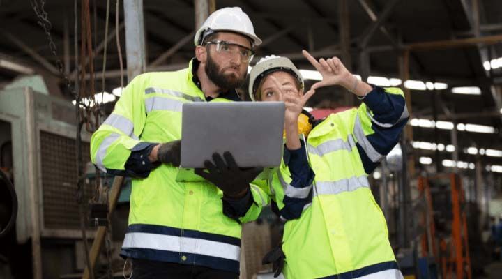 A man and a woman are standing next to each other in a factory looking at a laptop.