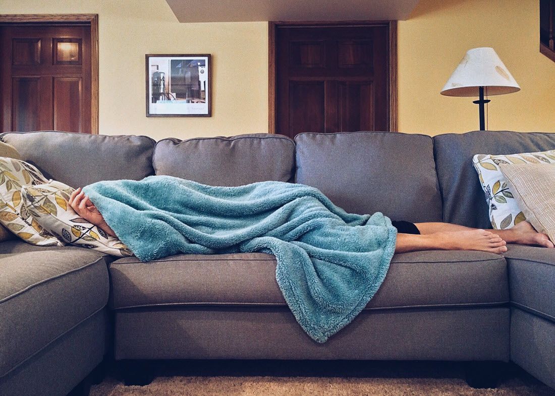 Person lying face down on a gray couch under a teal blanket in a living room.