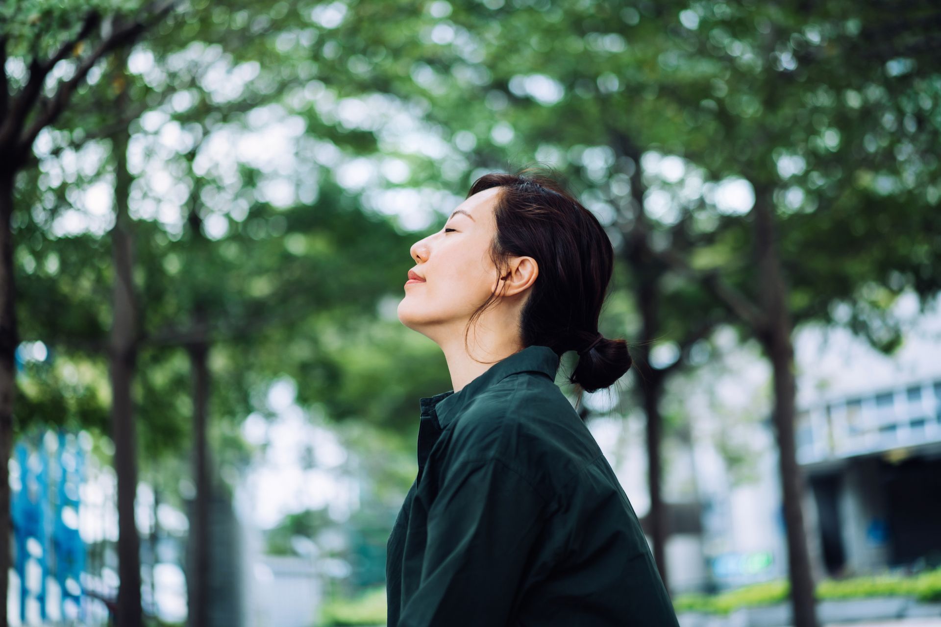 A woman in a green shirt is standing in a park with her eyes closed.