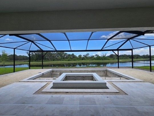 Empty rectangular pool under construction with stone coping and tiled deck, enclosed by a black screened lanai overlooking a lake and green landscape under a blue sky.