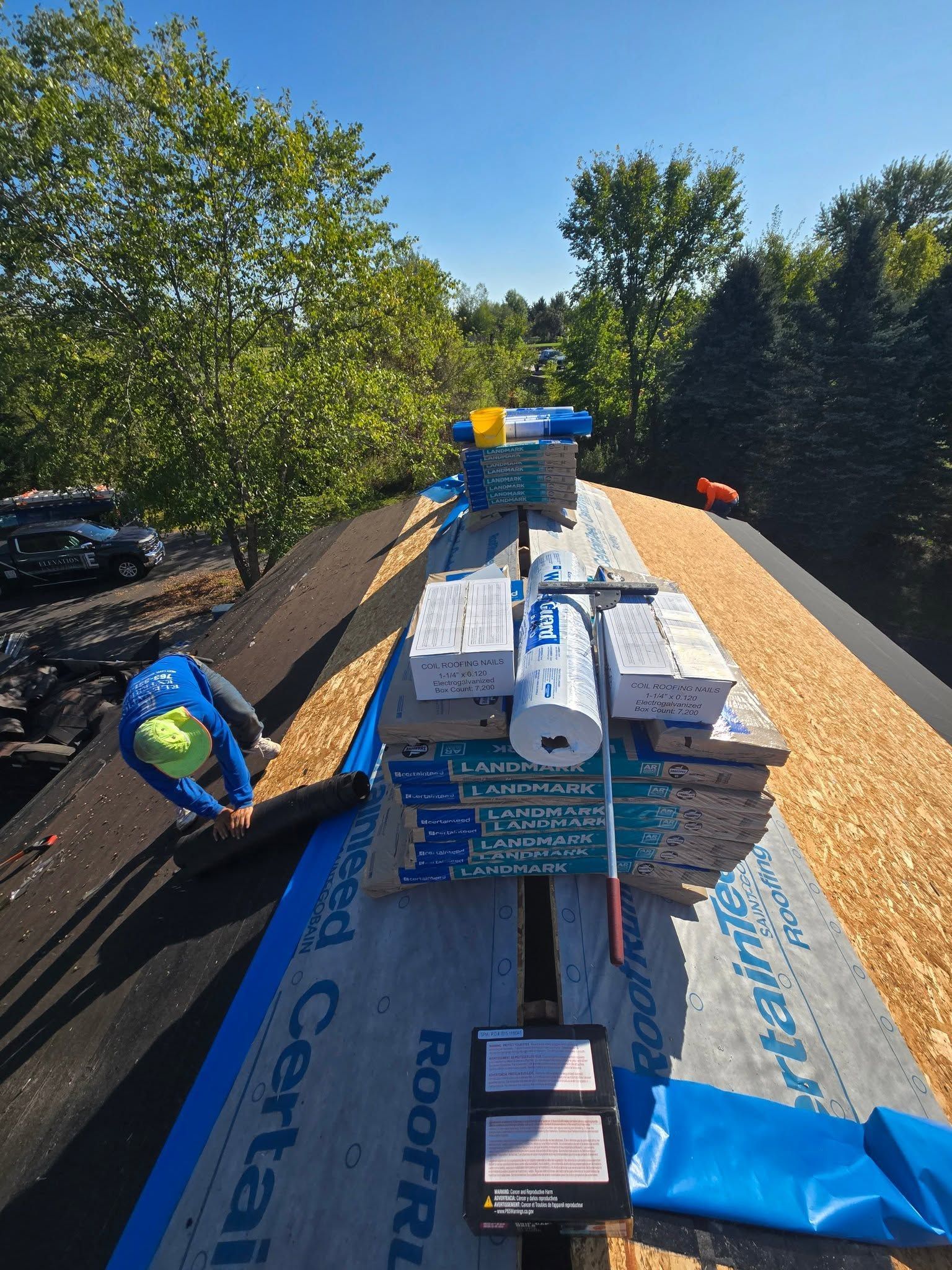 Roofer on a rooftop with new plywood and roofing materials. Sunny day with trees in the background.