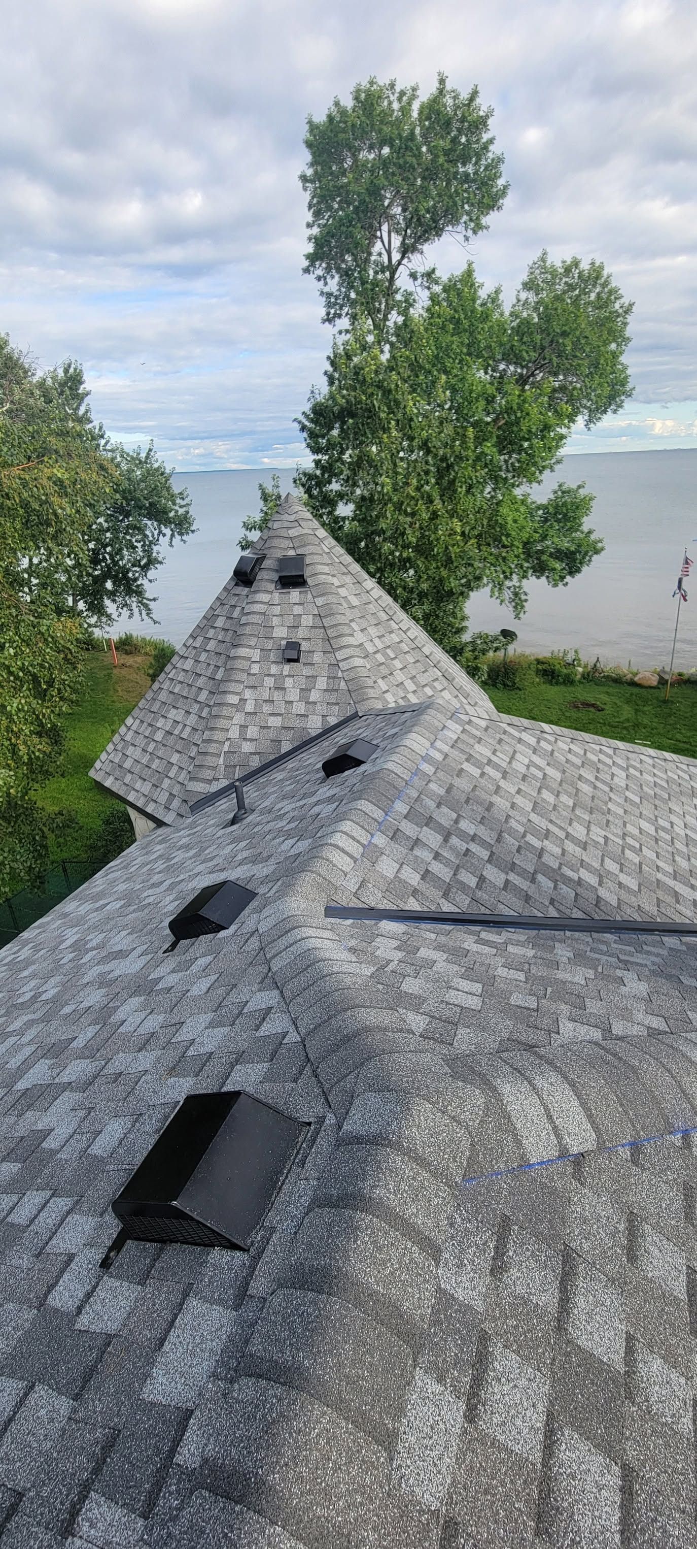 Gray shingled roof with multiple vent openings, overlooking a body of water and trees on a cloudy day.