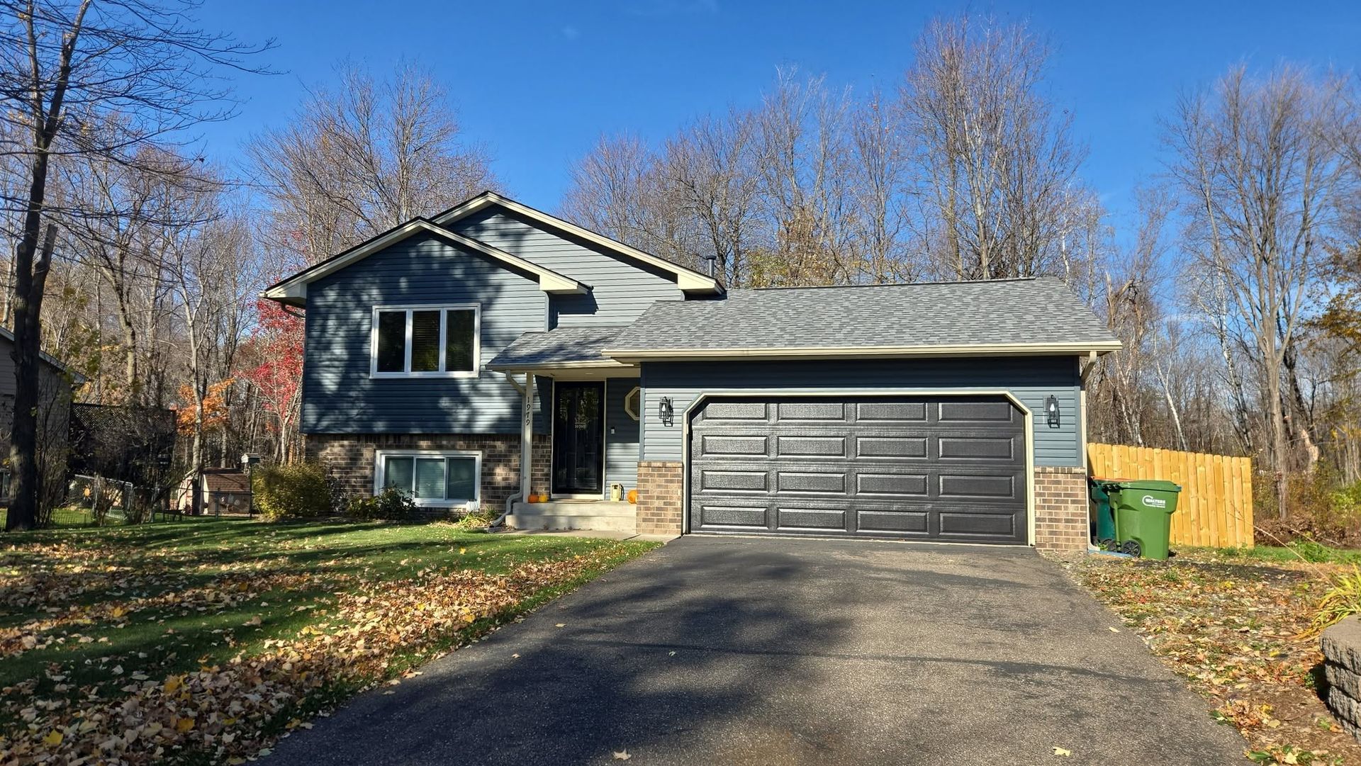 Blue house with gray garage door, driveway, green lawn, and fall foliage.