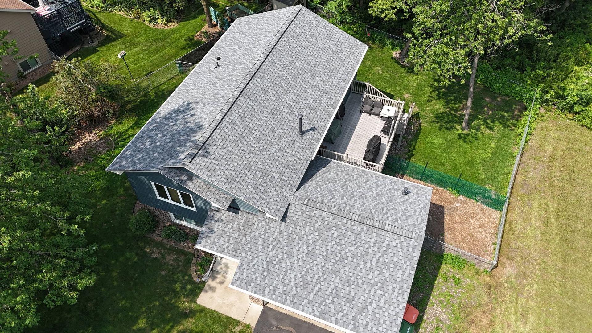 Aerial view of a gray-roofed house with a deck surrounded by trees and a green lawn.