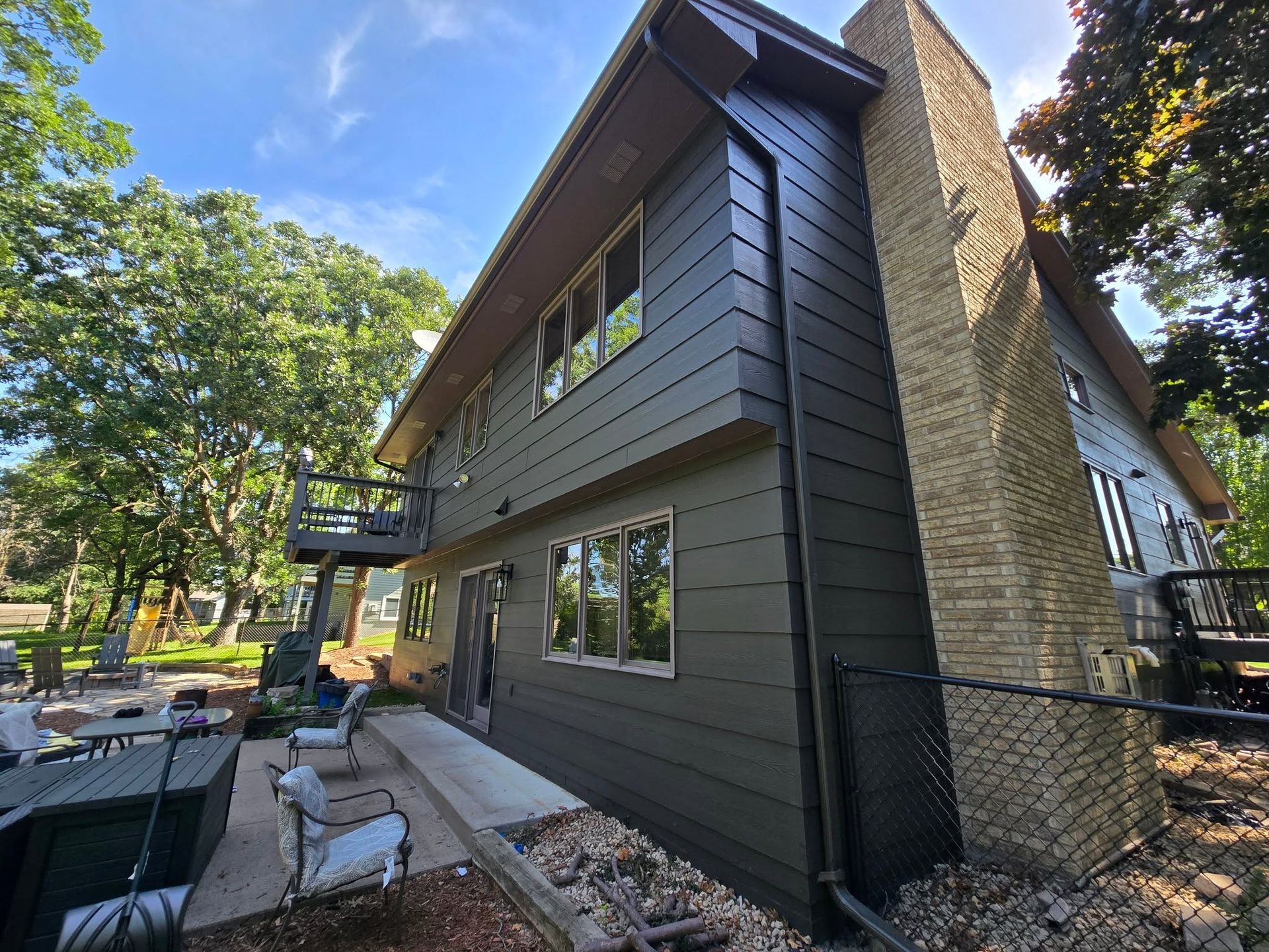 Two-story house with dark gray siding, a brick chimney, and a deck. Trees surround the house.