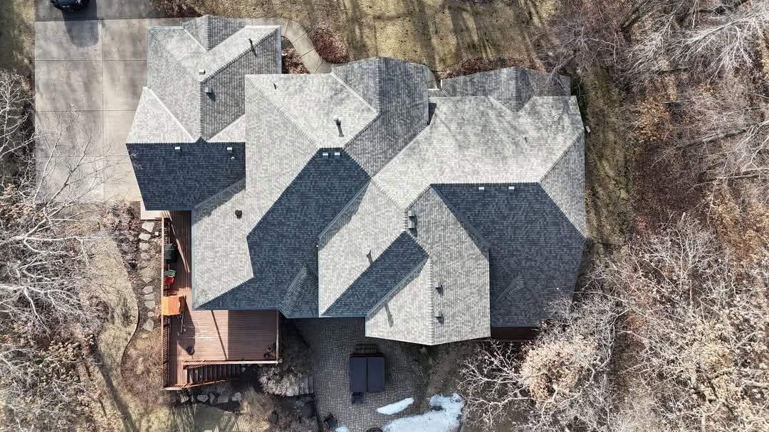 Aerial view of a complex gray-roofed house with a deck, surrounded by bare trees and brown foliage.