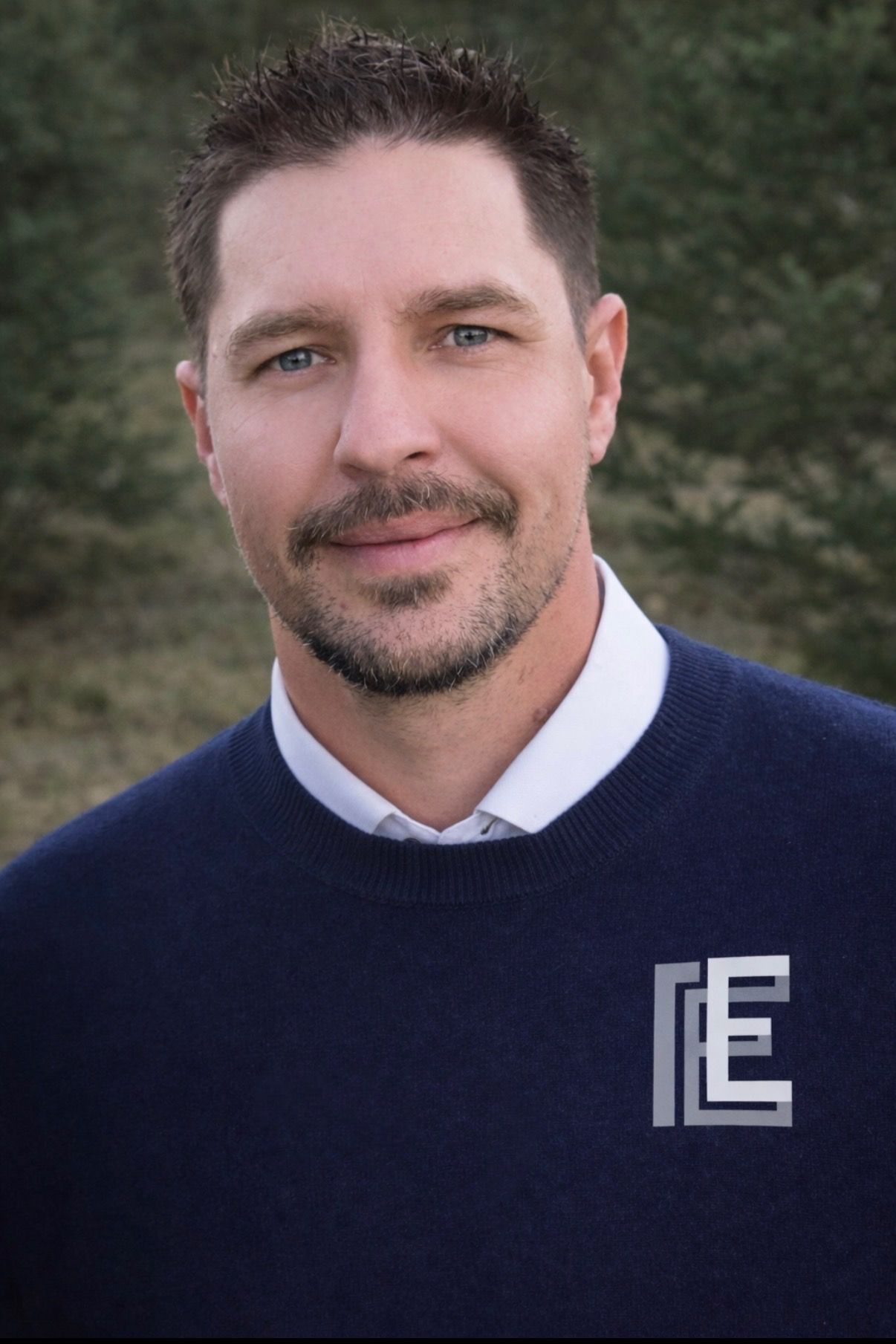 Man in black polo shirt with logo, smiling, in front of a white wall.