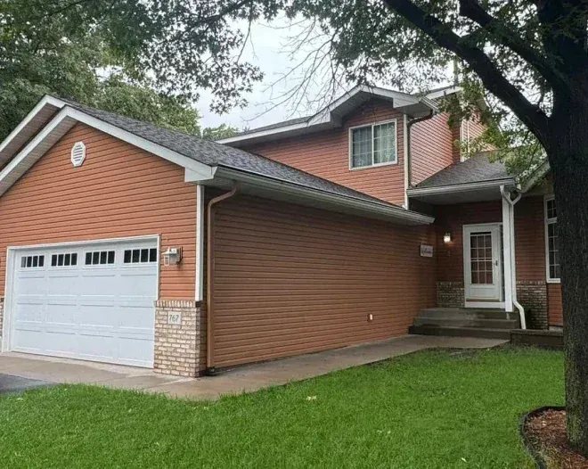 Two-story brown house with white garage door and entrance, on a green lawn with a tree.