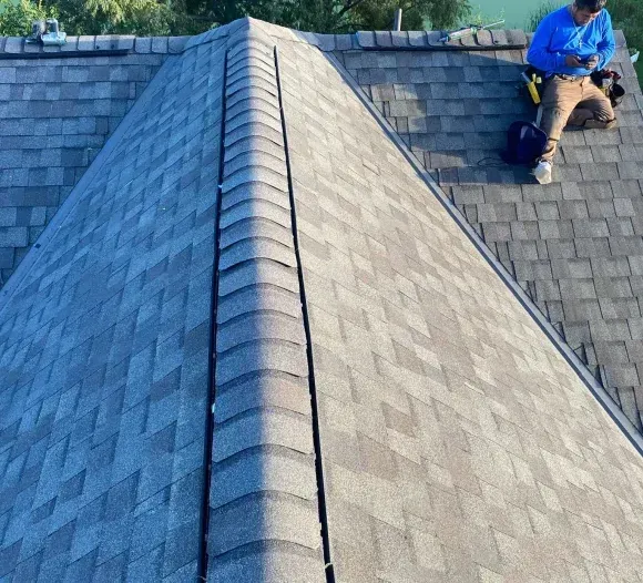 View of a house roof, asphalt shingles, with a person working on the edge.