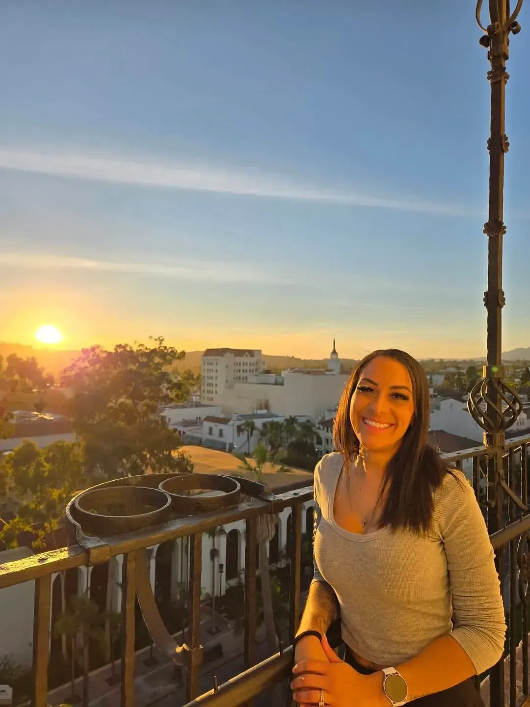 Woman on a balcony smiling at sunset over city buildings.