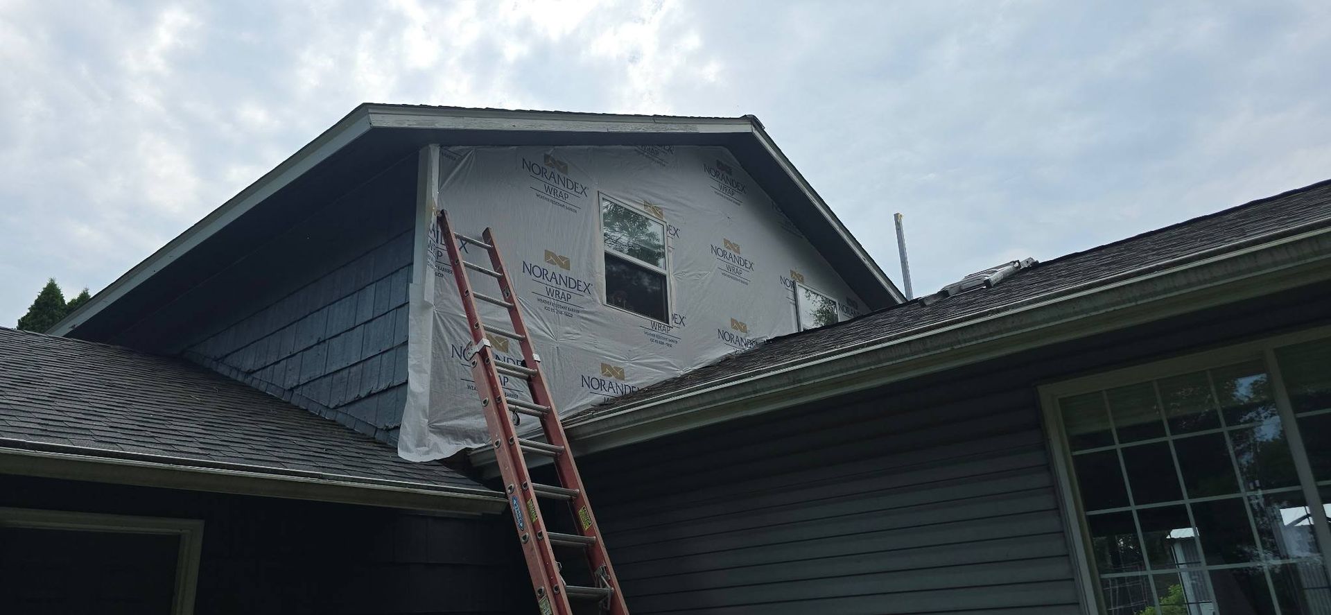 A house with siding partially covered in protective wrap, a ladder, and a cloudy sky.