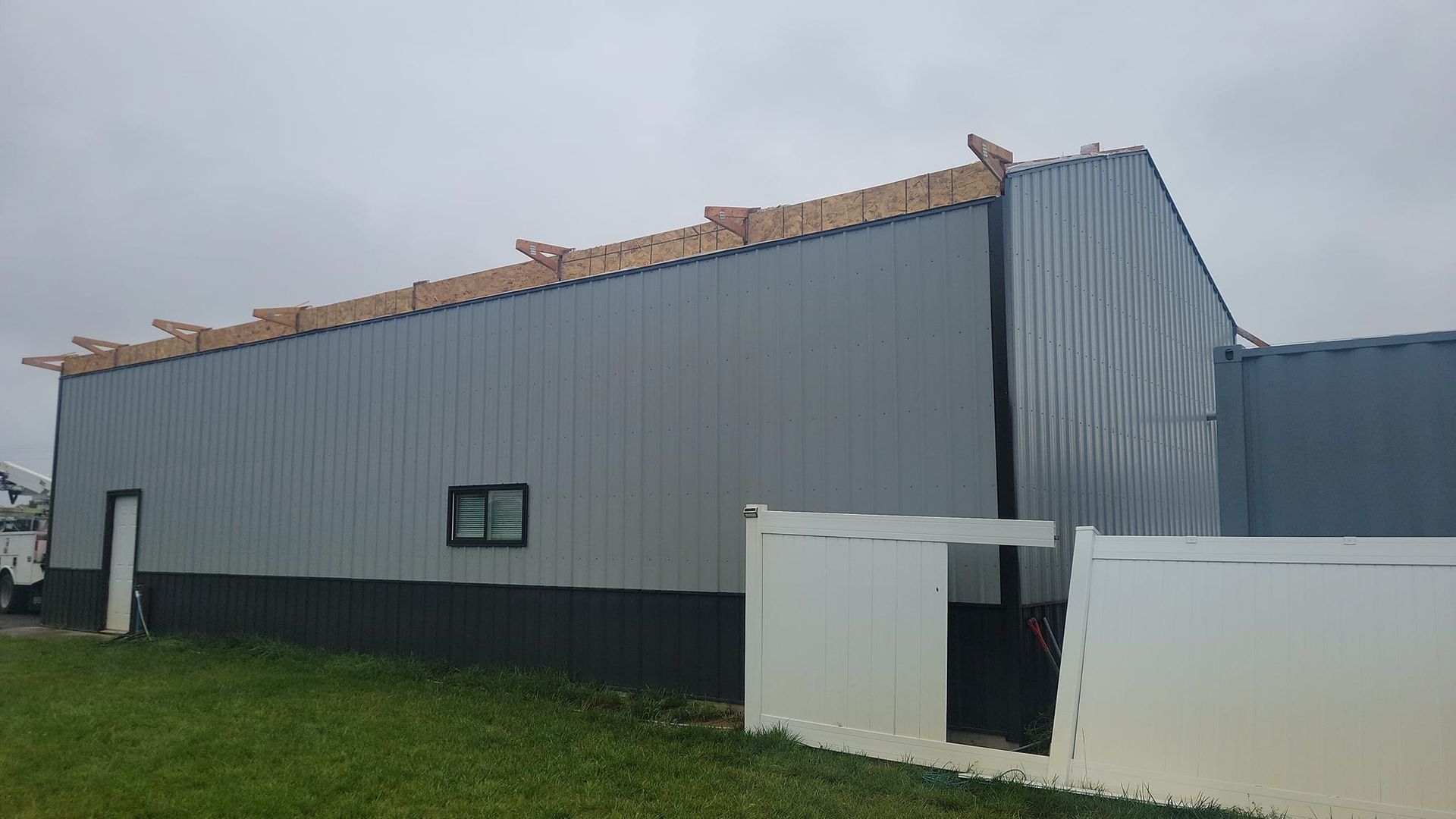 Gray metal building with exposed wooden roof framing, partially covered by tarp, next to white fence.