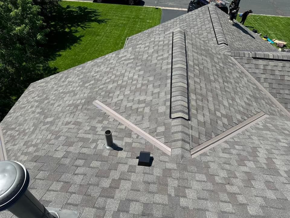 Gray asphalt shingle roof with vents and flashing. Workers on the roof. Green lawn in the background.
