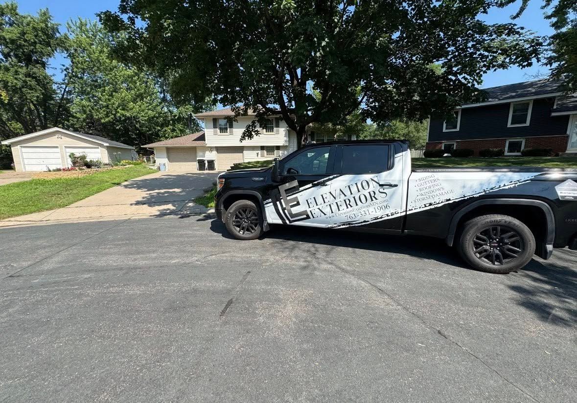 Black pickup truck parked in a driveway in front of a house. Trees and a sunny sky are visible.