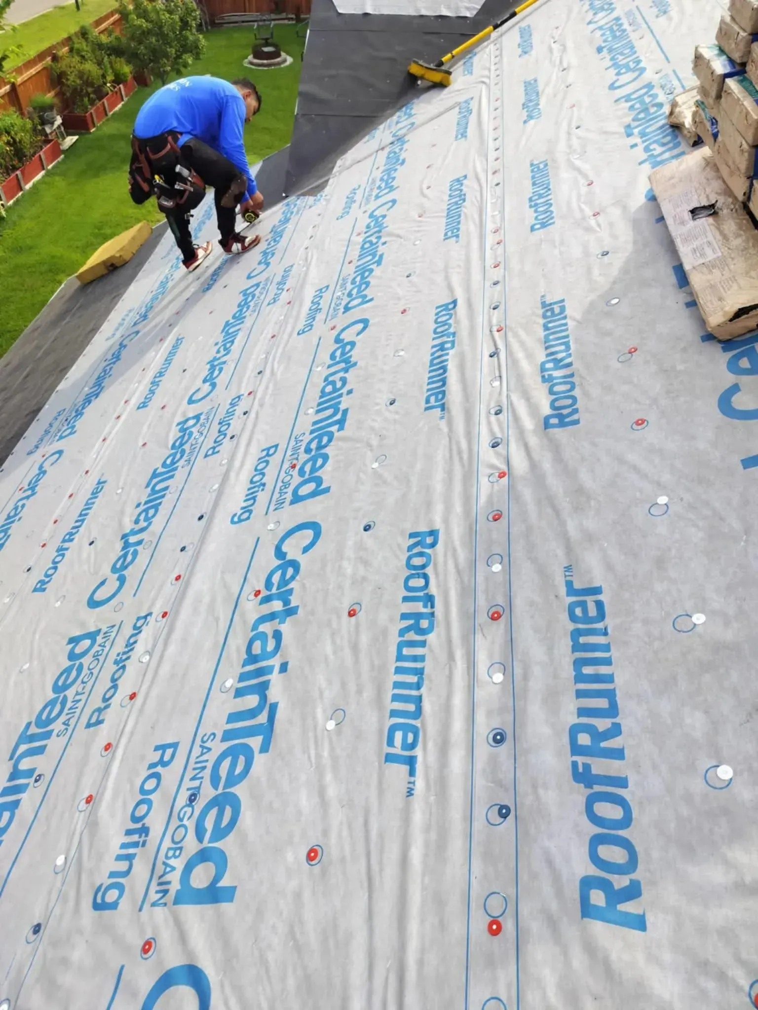 Roofer installing shingles on a roof covered with white underlayment, holding a tool. Green lawn in the background.