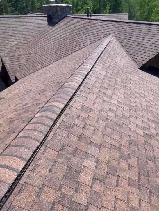 Brown asphalt shingle roof with a central ridge and chimney in the background.