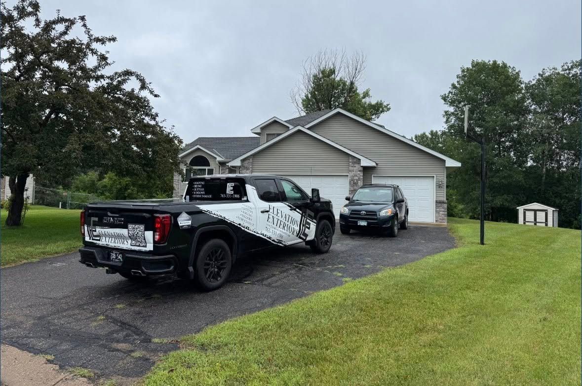 Black and white truck parked in front of a house on a driveway with a car, surrounded by green grass and trees.