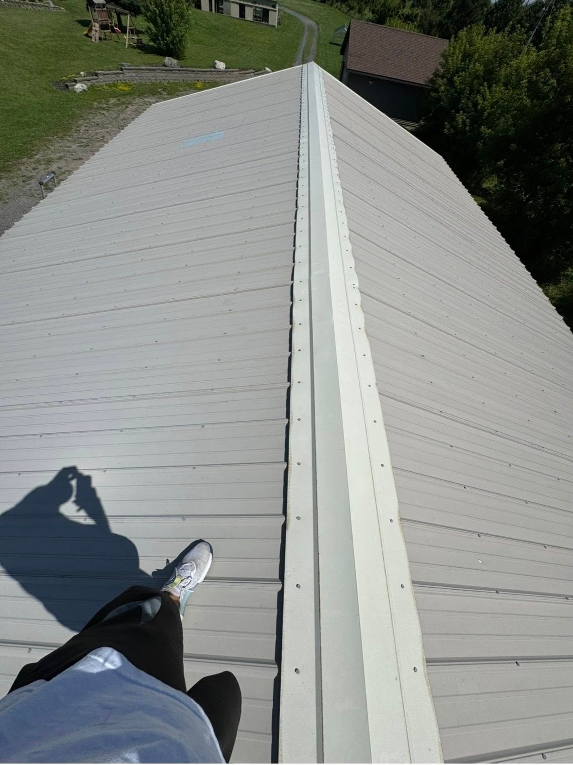 Person on metal roof, shadow visible, looking down. Gray roof panels, white trim, sunny outdoor setting.