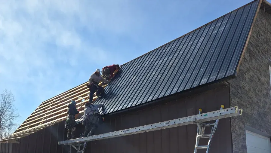 Construction workers installing a dark metal roof on a brown building under a partly cloudy sky.
