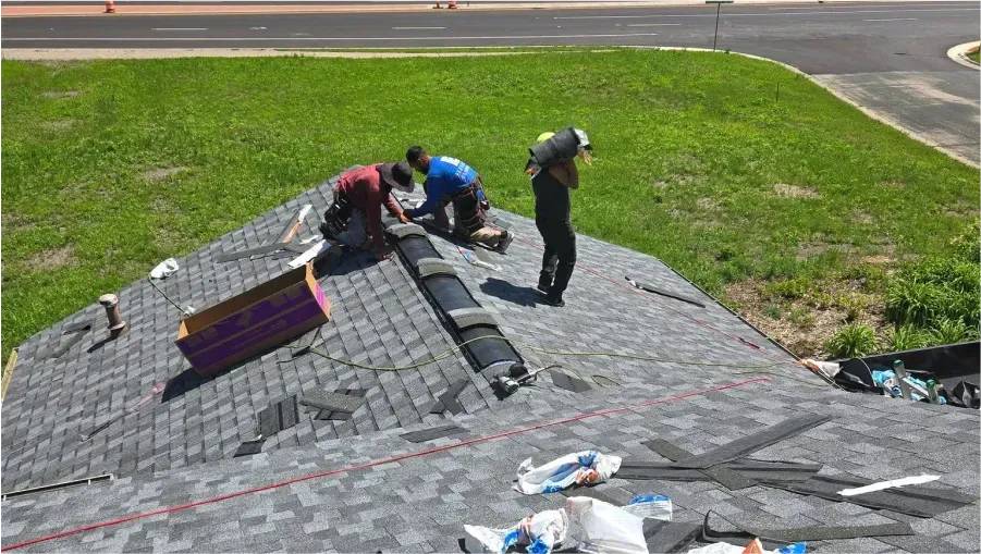 Three roofers installing shingles on a house roof next to a grassy area and road.