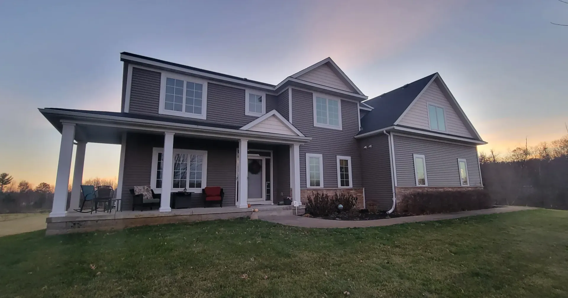 Two-story house with gray siding, white trim, and a porch, set in a grassy yard at dusk.