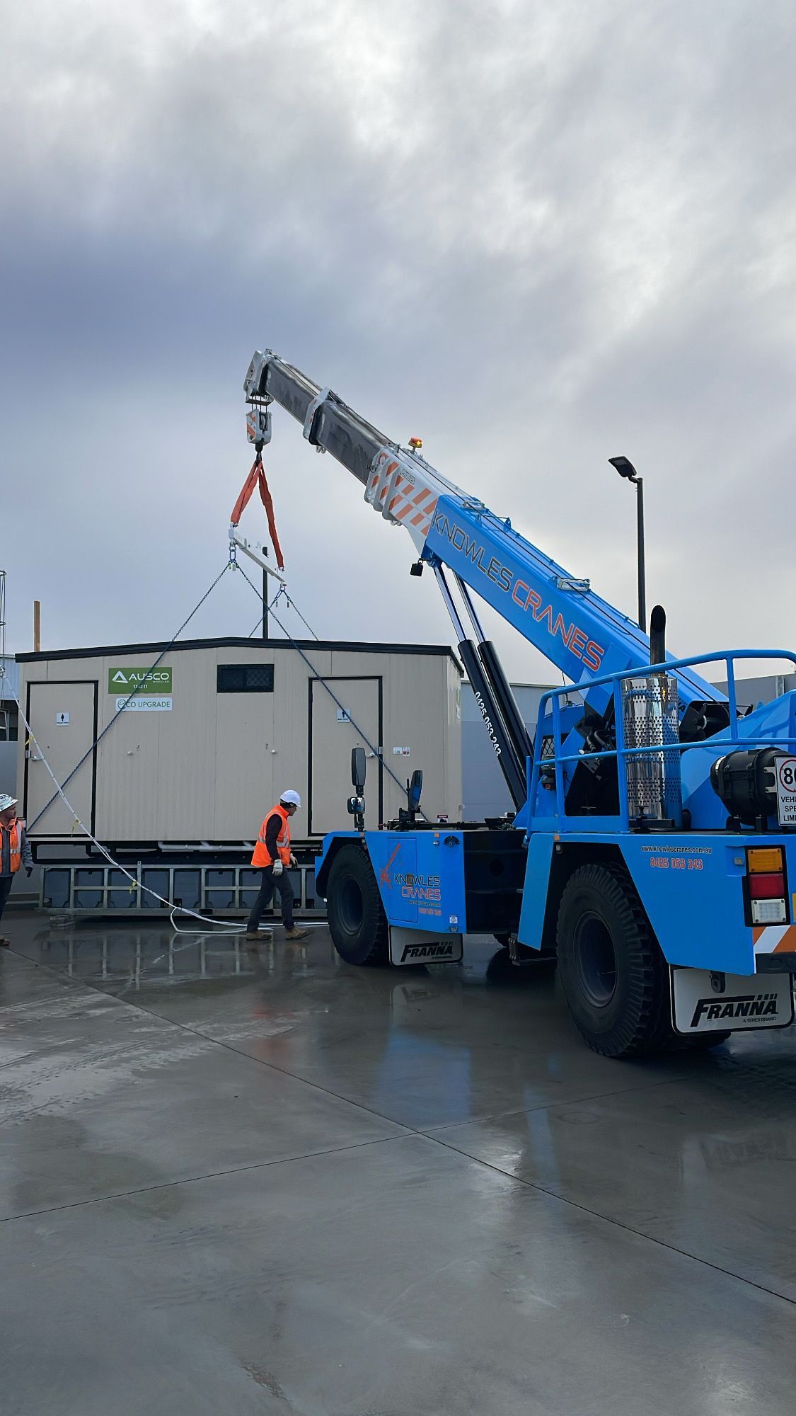 Knowles Cranes Franna crane lifting a portable site office while workers supervise the operation on a wet jobsite.