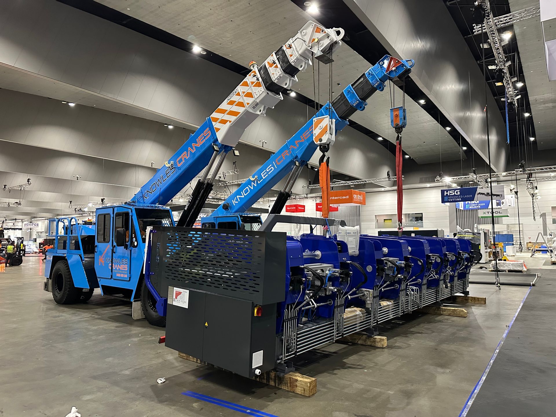 Dual Knowles Cranes lifting industrial machinery at indoor exhibition setup with signage in background.