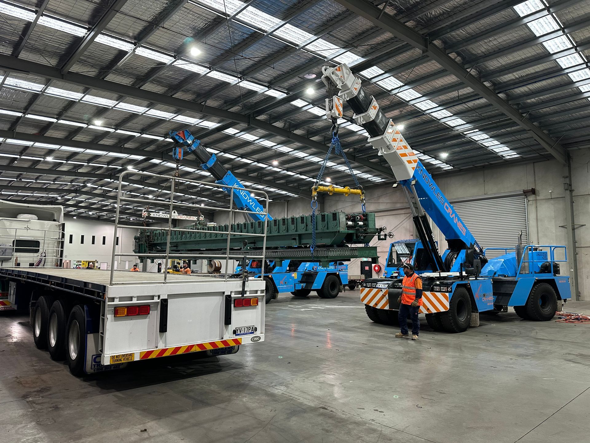 Twin Knowles Cranes Franna units lifting a large industrial machine onto a flatbed truck inside a warehouse.