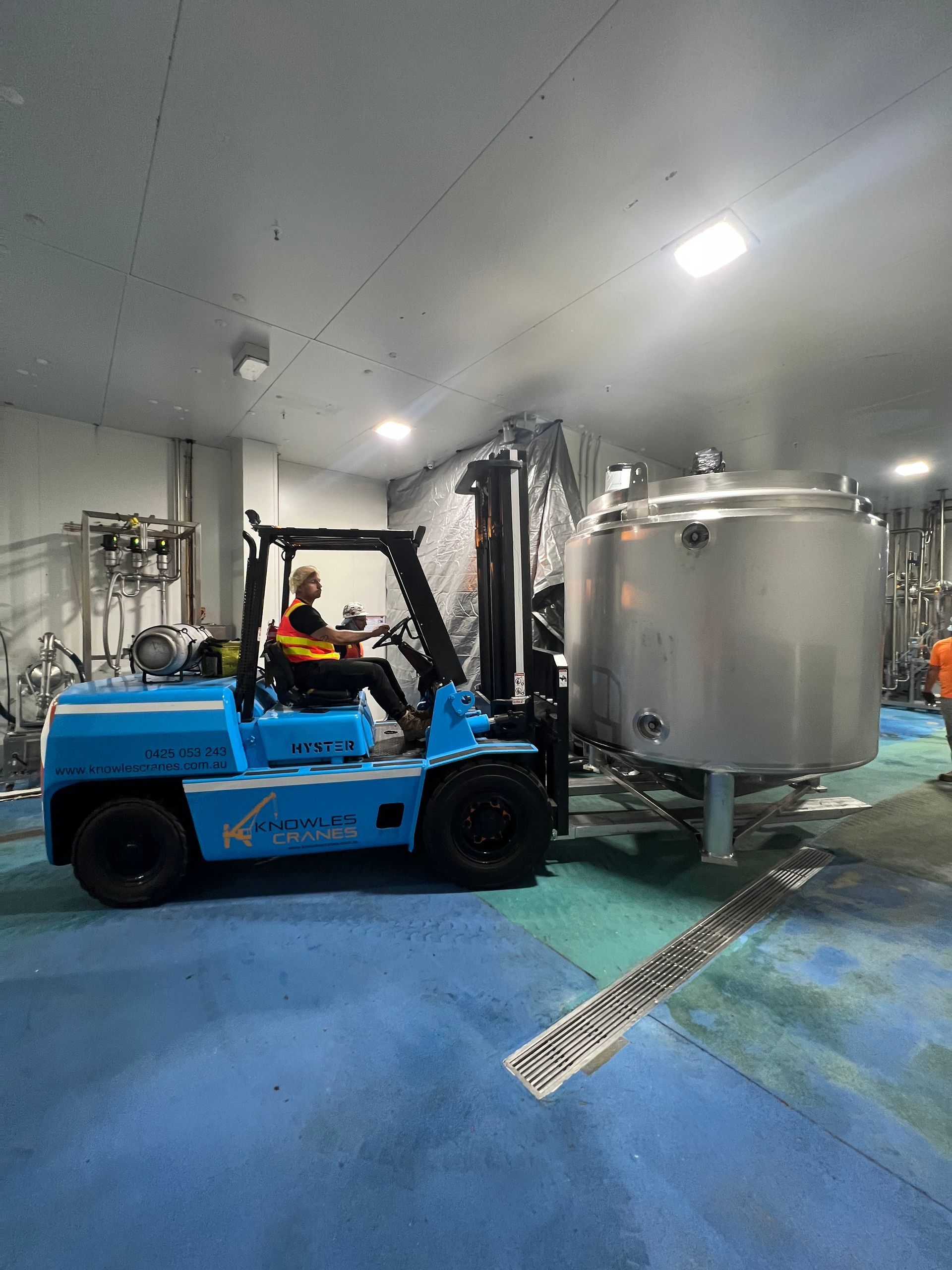 A blue forklift is moving a large stainless steel tank in a factory.