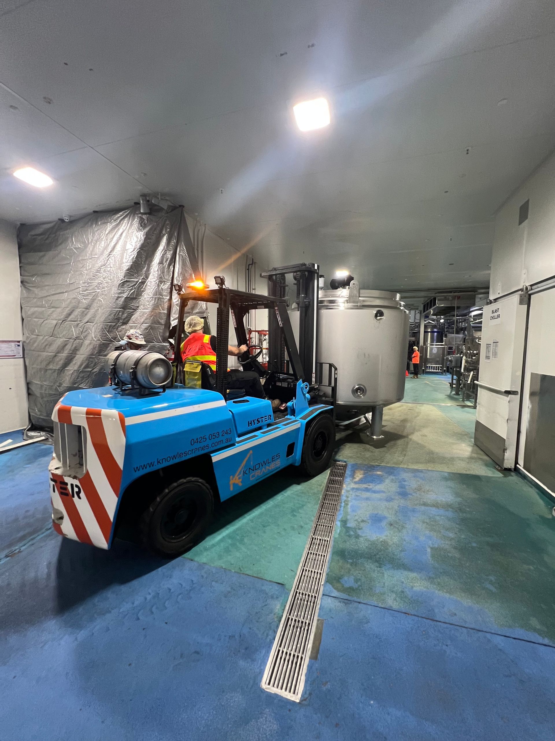 Forklift moving stainless steel tank inside facility with workers in safety gear