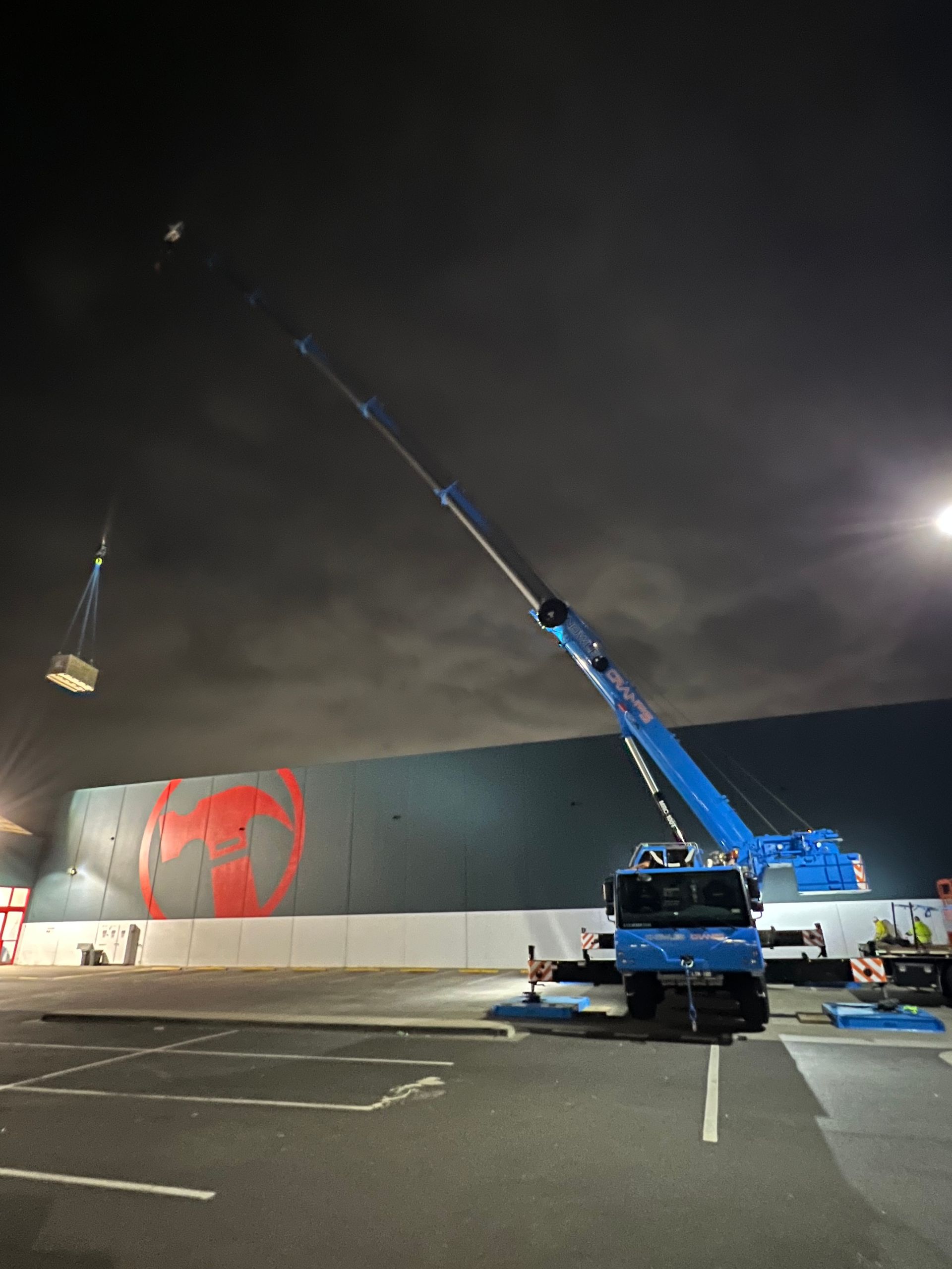 Crane operating at night in a parking lot, lifting a load under bright floodlights.