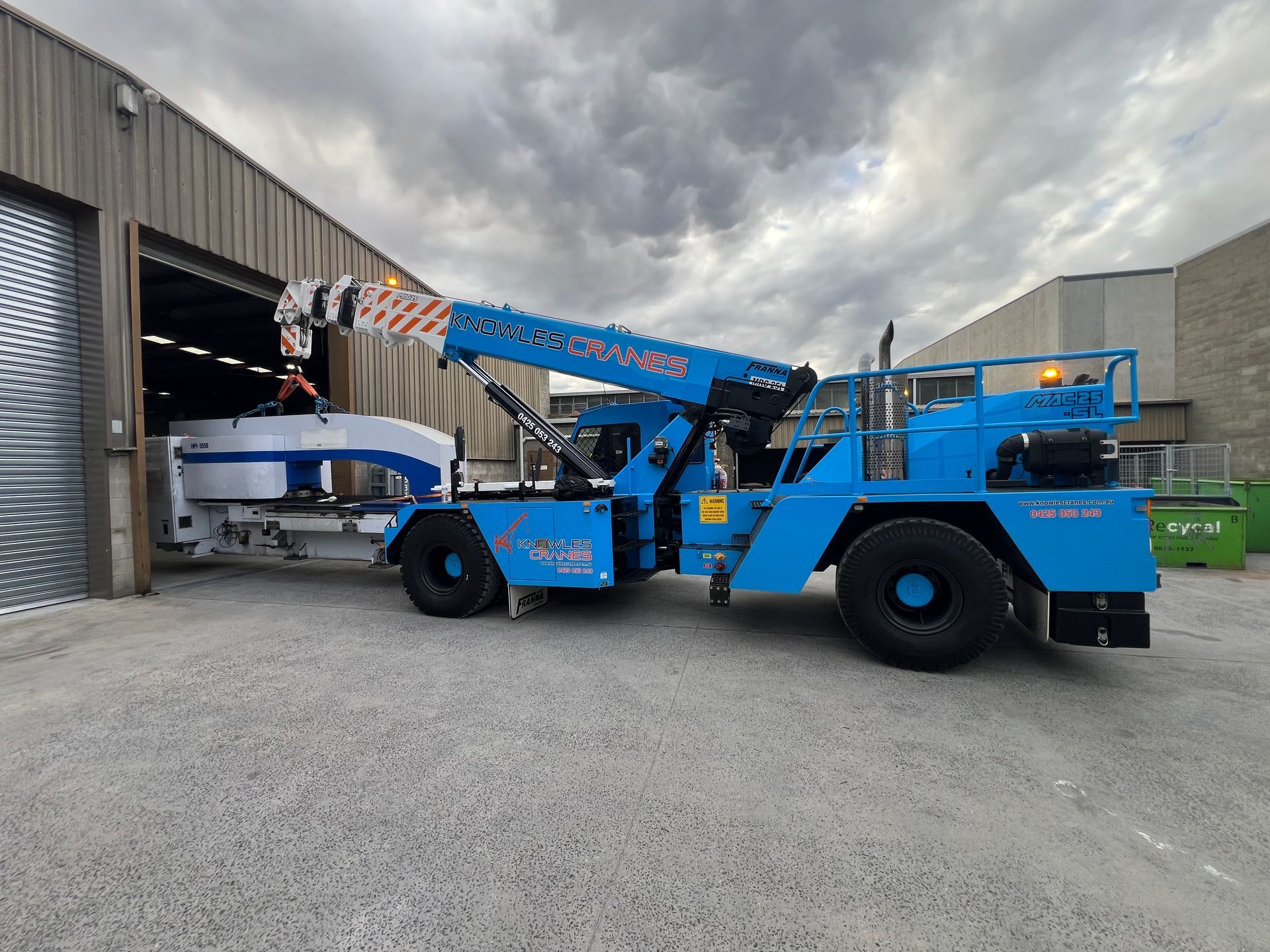 Franna crane extracting heavy machinery from a factory under cloudy skies.