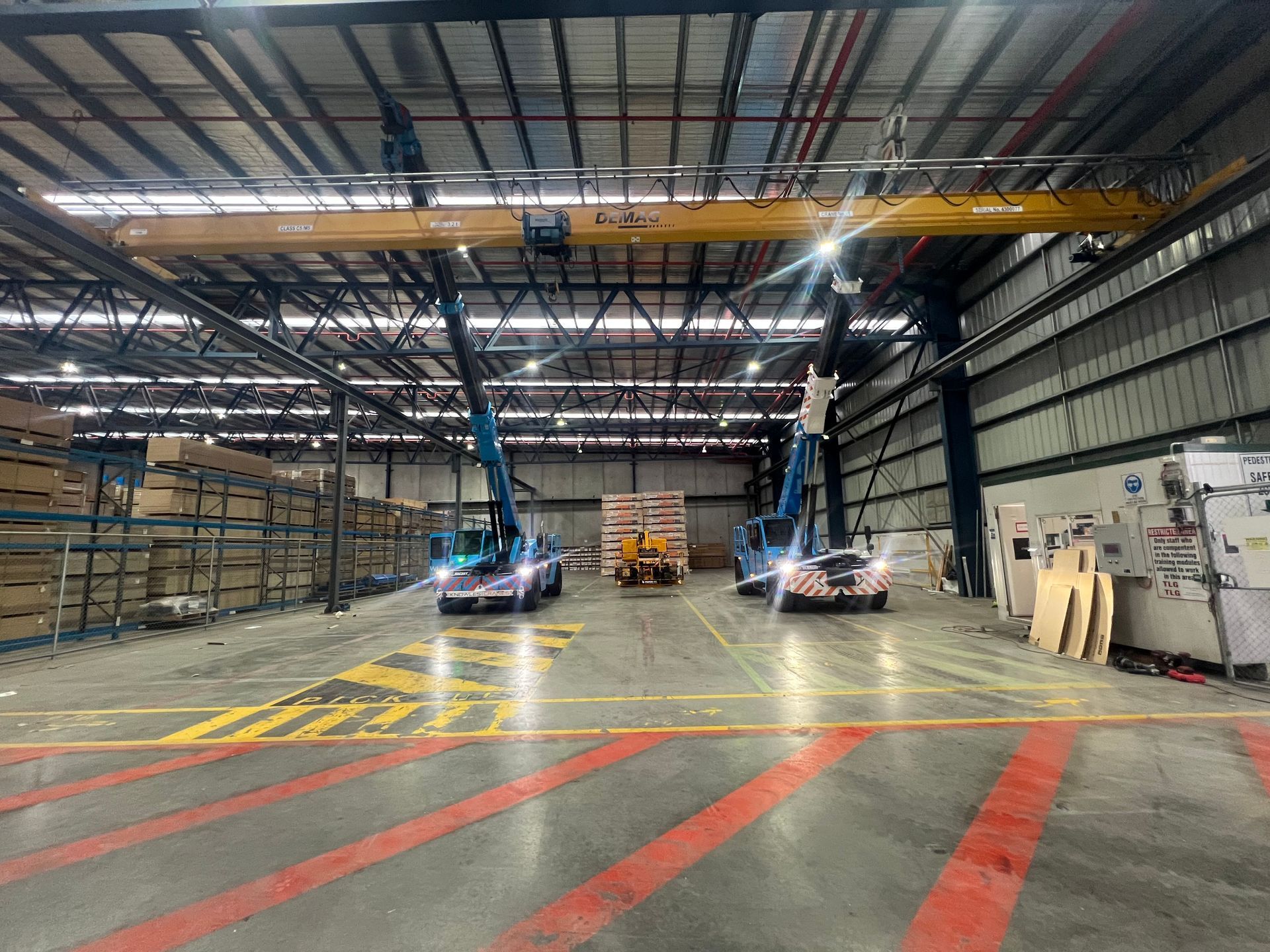 Two Knowles Cranes Franna units are performing a dual lift inside a large warehouse with an overhead crane system.