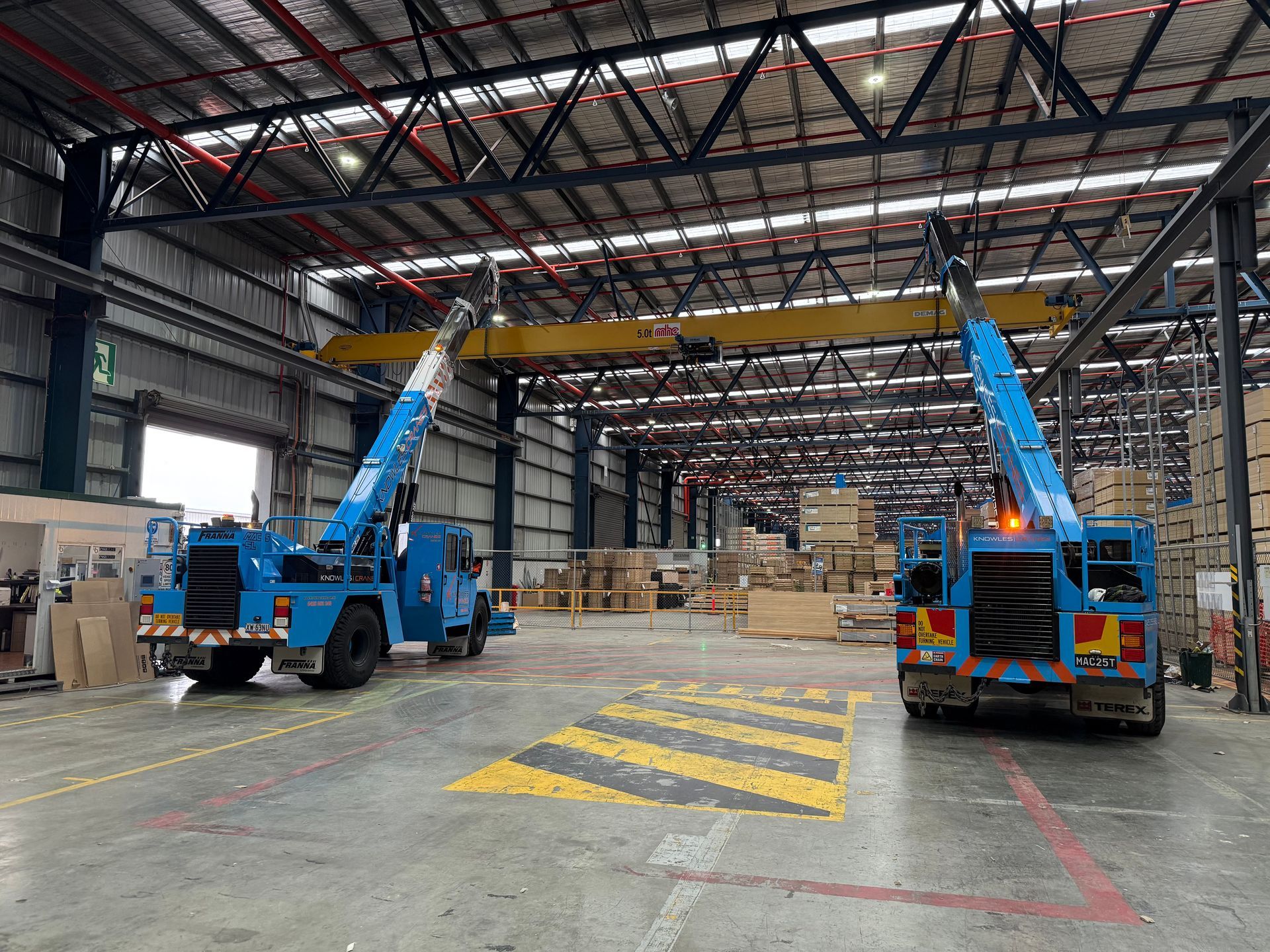 Rear view of two Knowles Cranes Franna units positioned inside a warehouse surrounded by timber stacks and racking.