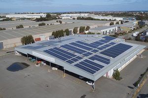 Solar panels on a large industrial building roof. Gray and blue panels against a gray roof, in a sunny outdoor setting.