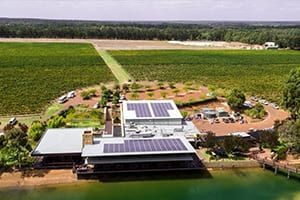 Winery with solar panels on the roof, next to a vineyard and lake.