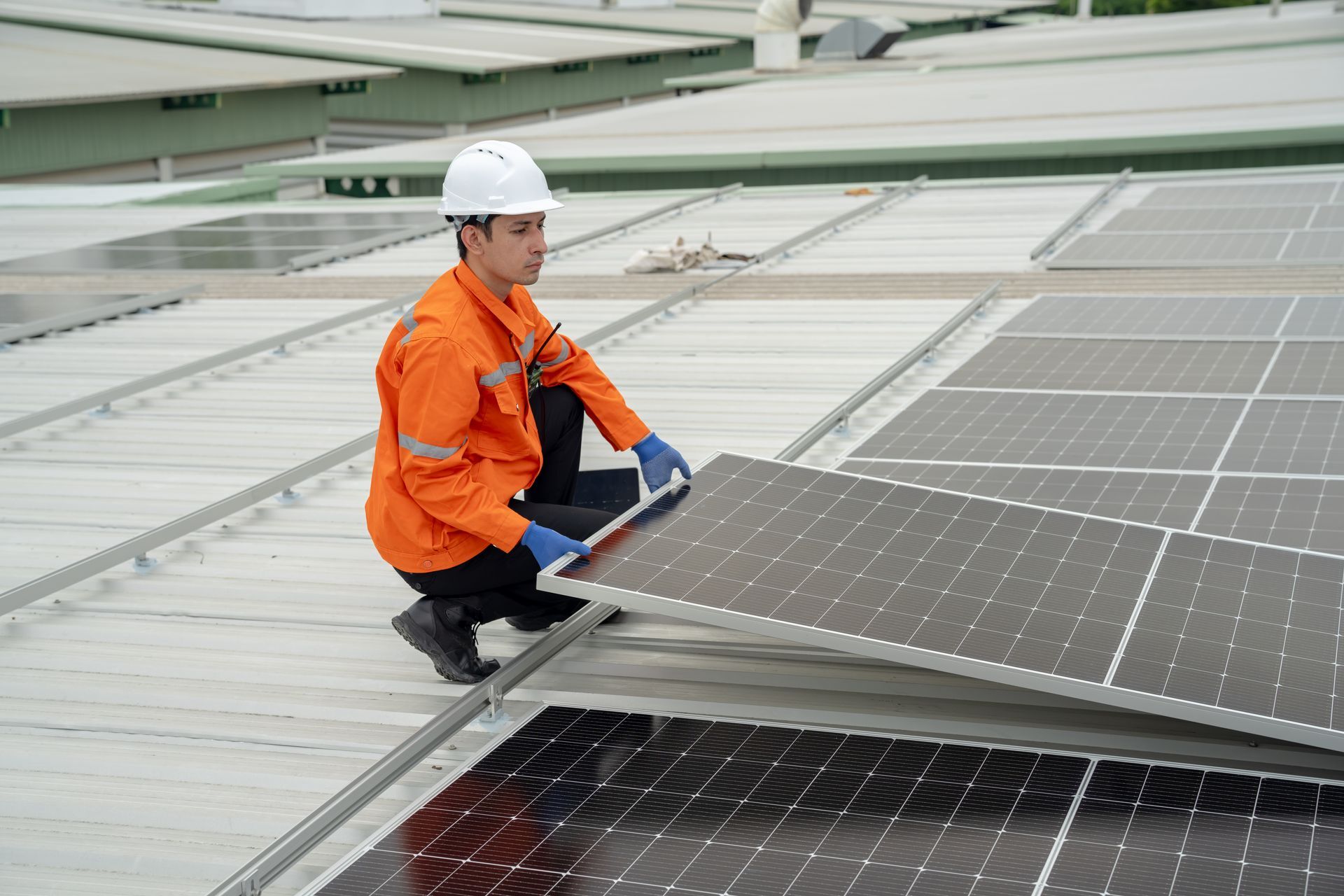 Worker in orange suit installing solar panel on a rooftop, wearing a white hard hat.