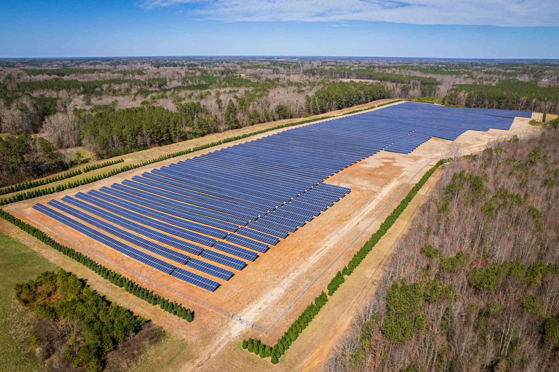 Aerial view of a large solar panel farm in a field, surrounded by trees, under a blue sky.