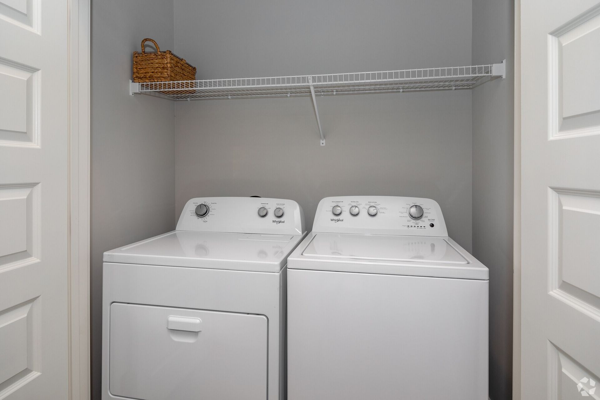 Laundry closet with white washer and dryer side by side under a wire shelf and a basket.