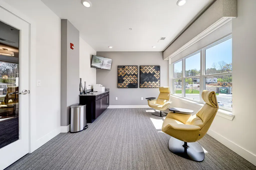 Interior community lounge with large windows, two yellow lounge chairs, and a small table.