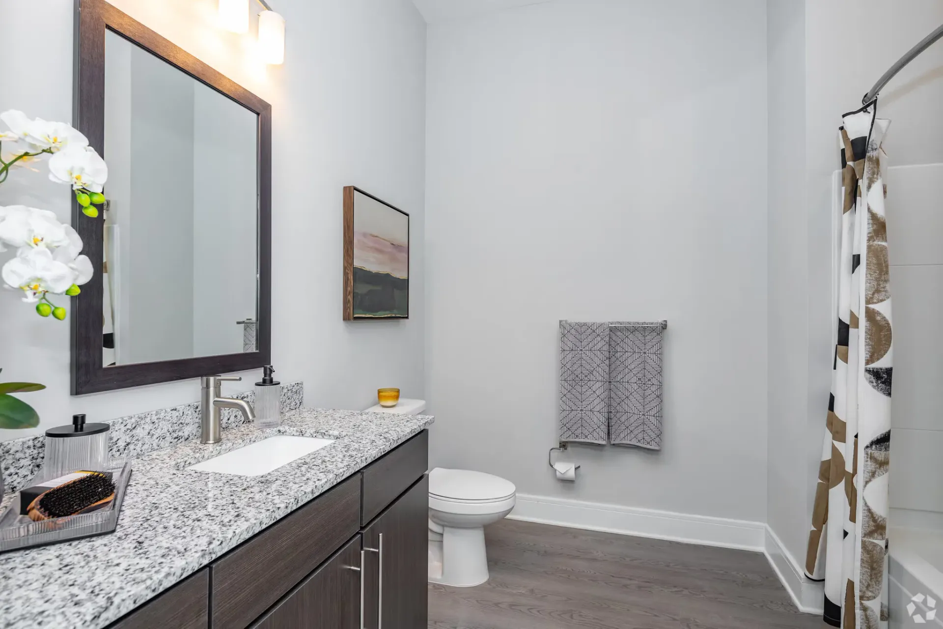 Bathroom interior with granite countertop, dark wood vanity, large mirror, toilet, and a patterned shower curtain.