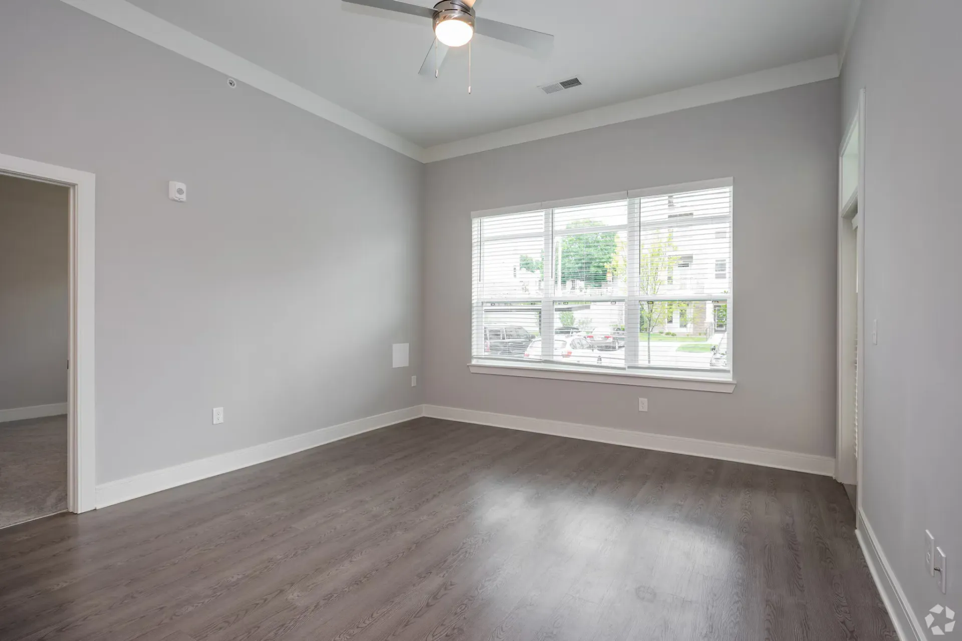 Empty living area in an apartment with a large window, gray walls, and a ceiling fan.